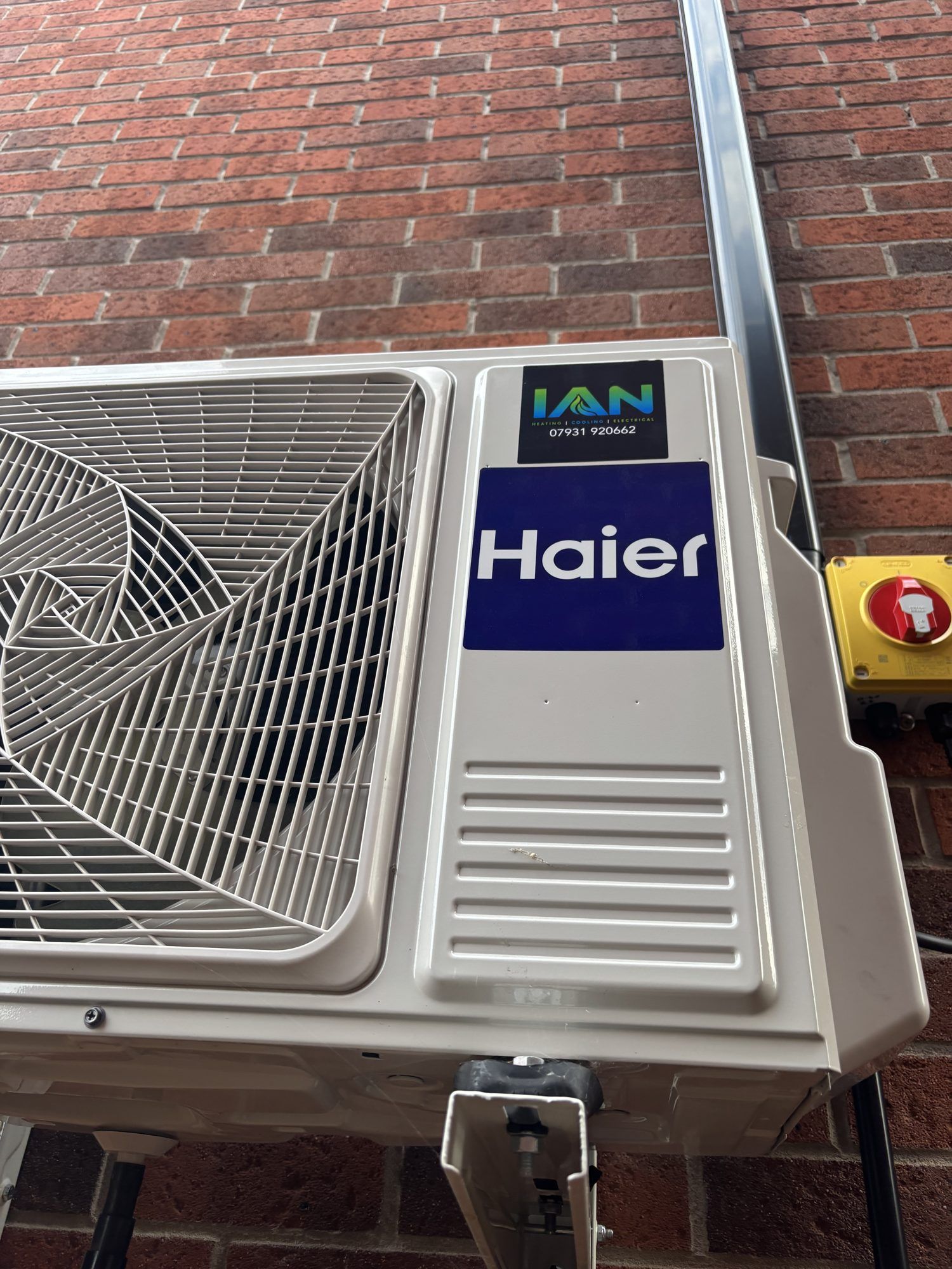 The outdoor unit of a Haier air conditioner on wall brackets against a brick wall, with a yellow isolator switch beside it