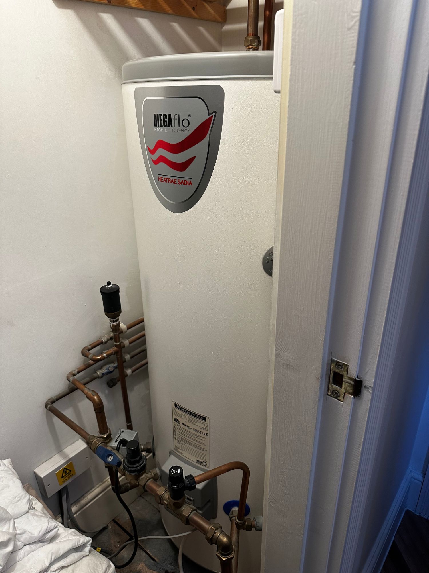 A white Heatrae Sadia Megaflo High Efficiency hot water cylinder in an airing cupboard, with copper pipework and valves beside it and an electrical spur on the wall below