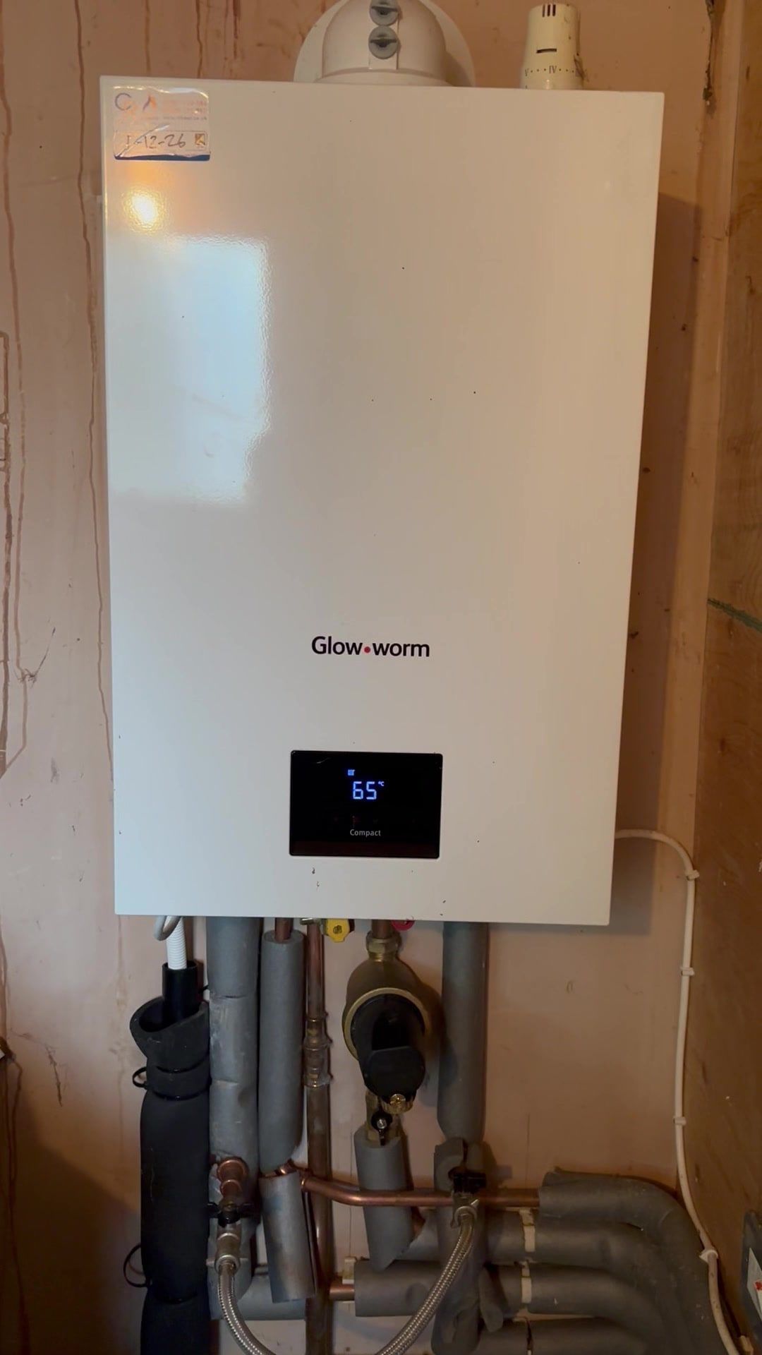 A white wall-hung Glow-worm combi boiler with its display showing 65 degrees, insulated pipework and a filter below it