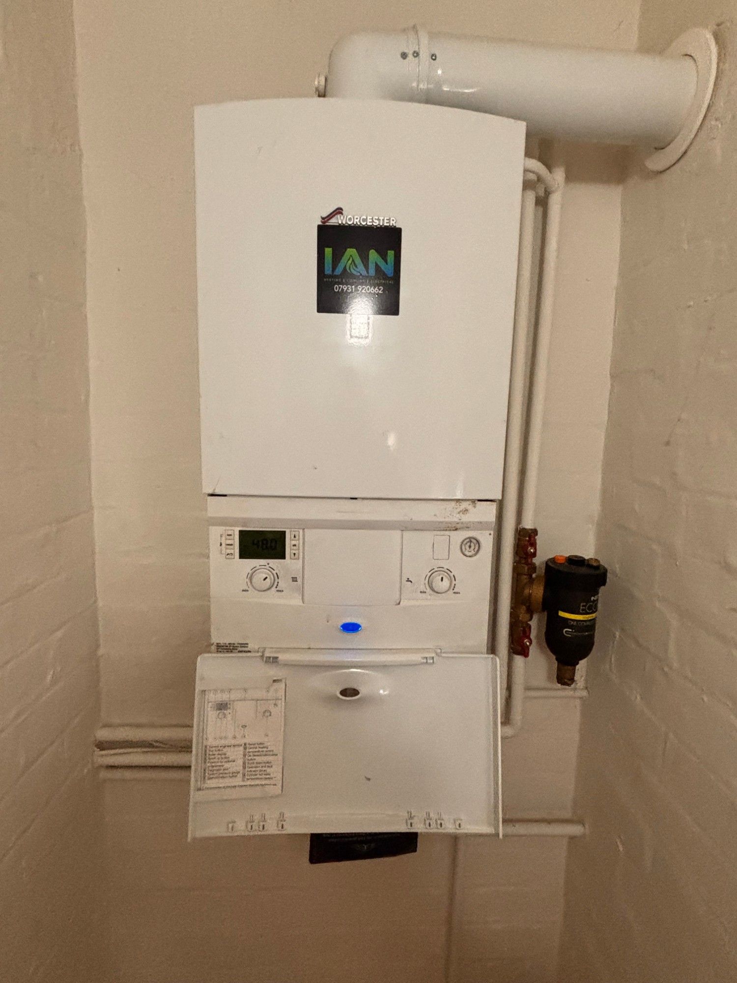 A white wall-hung Worcester Bosch gas boiler with Ian's sticker on the front, its control flap open and the display showing 48 degrees, under a white flue pipe with a black filter on the pipework beside it