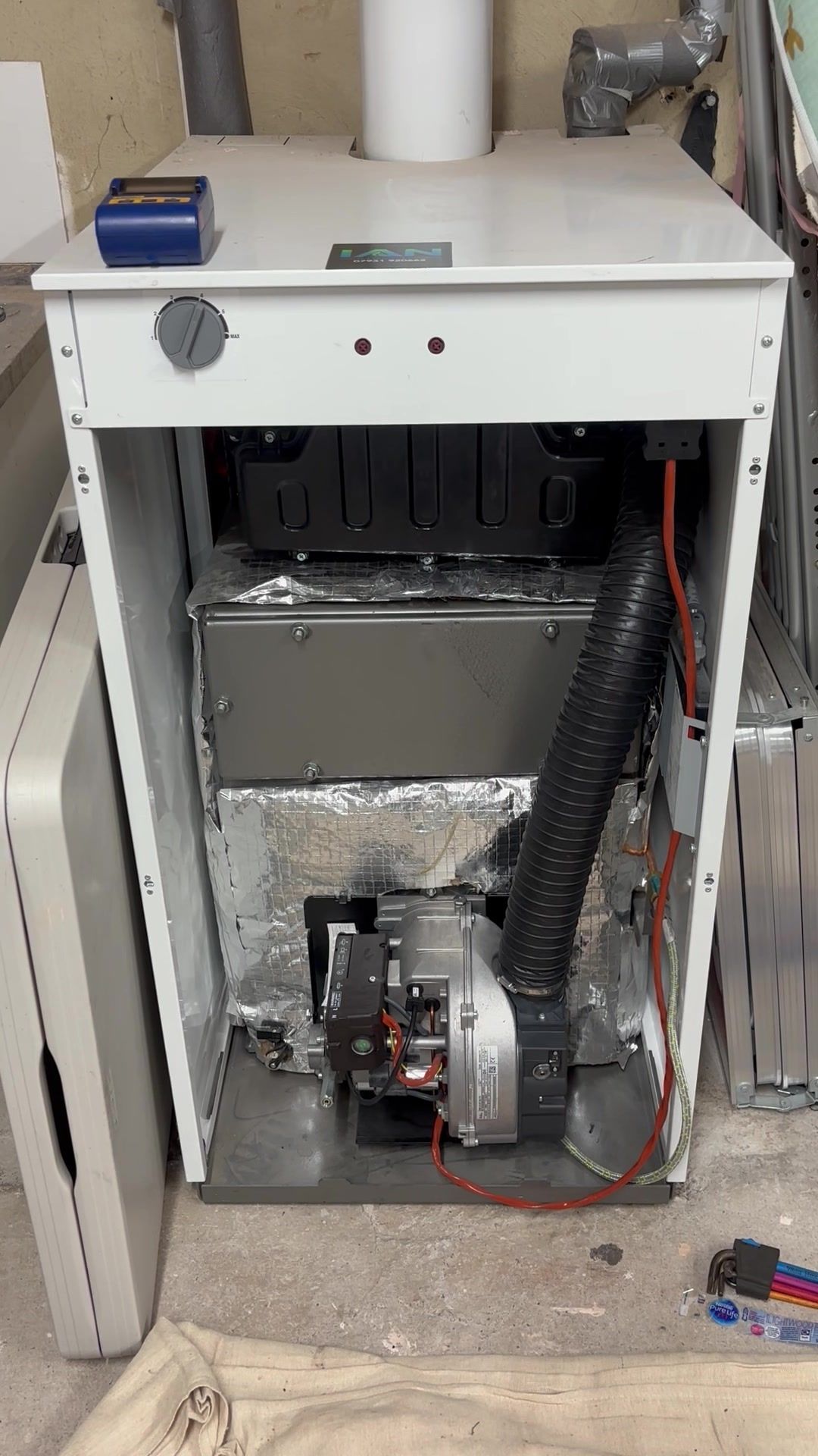 A white floor-standing oil boiler with its front panel off, showing the burner and its black air hose at the bottom, with Ian's sticker on the top and a dust sheet on the floor