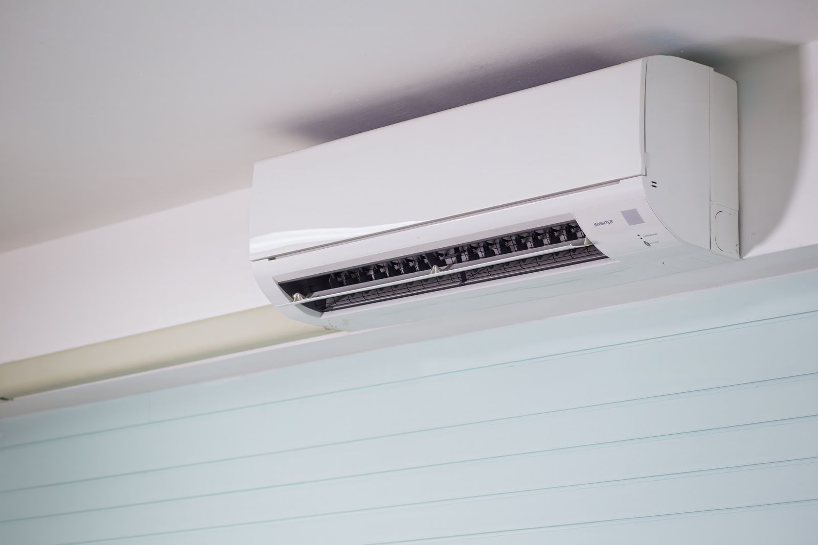 A white wall-mounted air conditioning unit near the ceiling of a room with a pale blue panelled wall