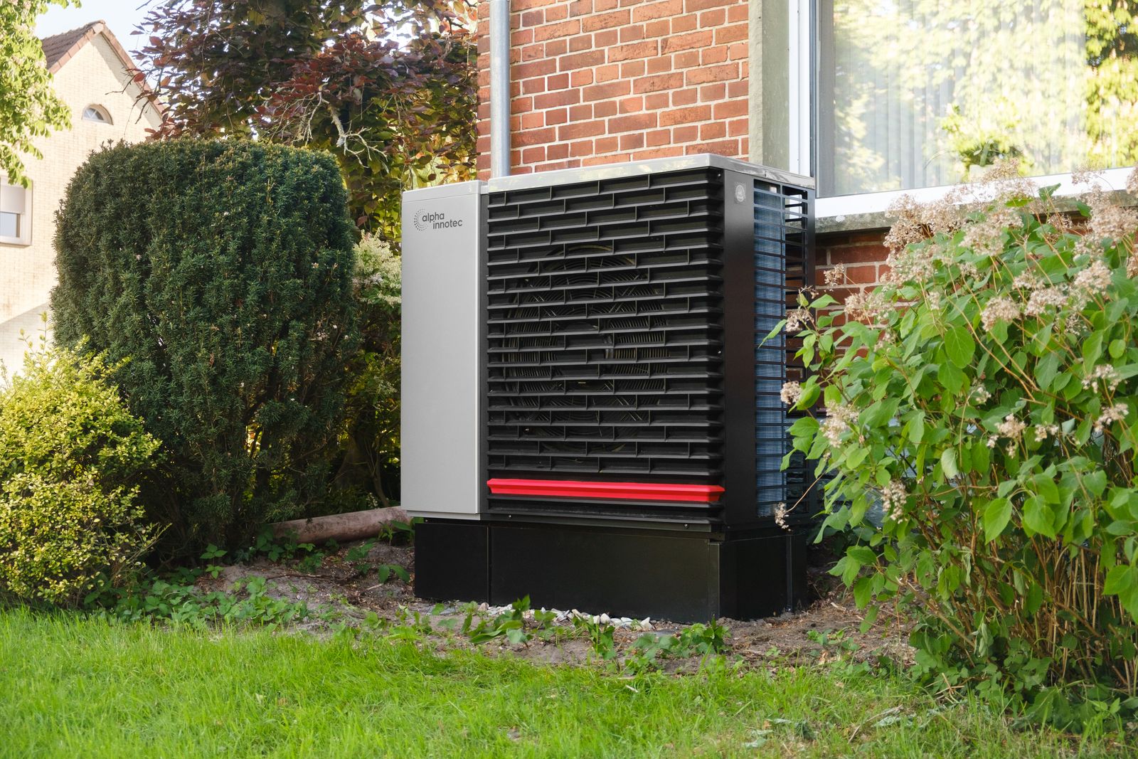 A black and grey air source heat pump on a concrete base against the brick wall of a house, with shrubs either side