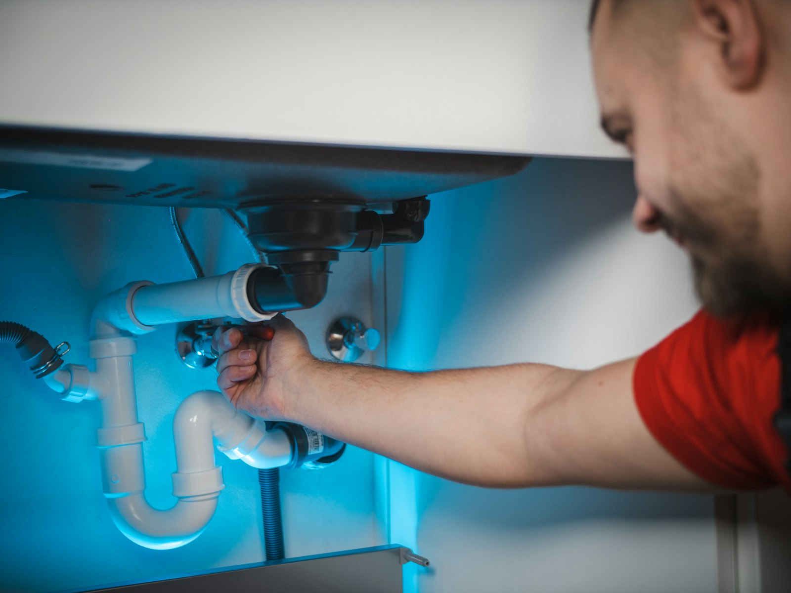 A plumber in a red T-shirt working on the pipework under a kitchen sink