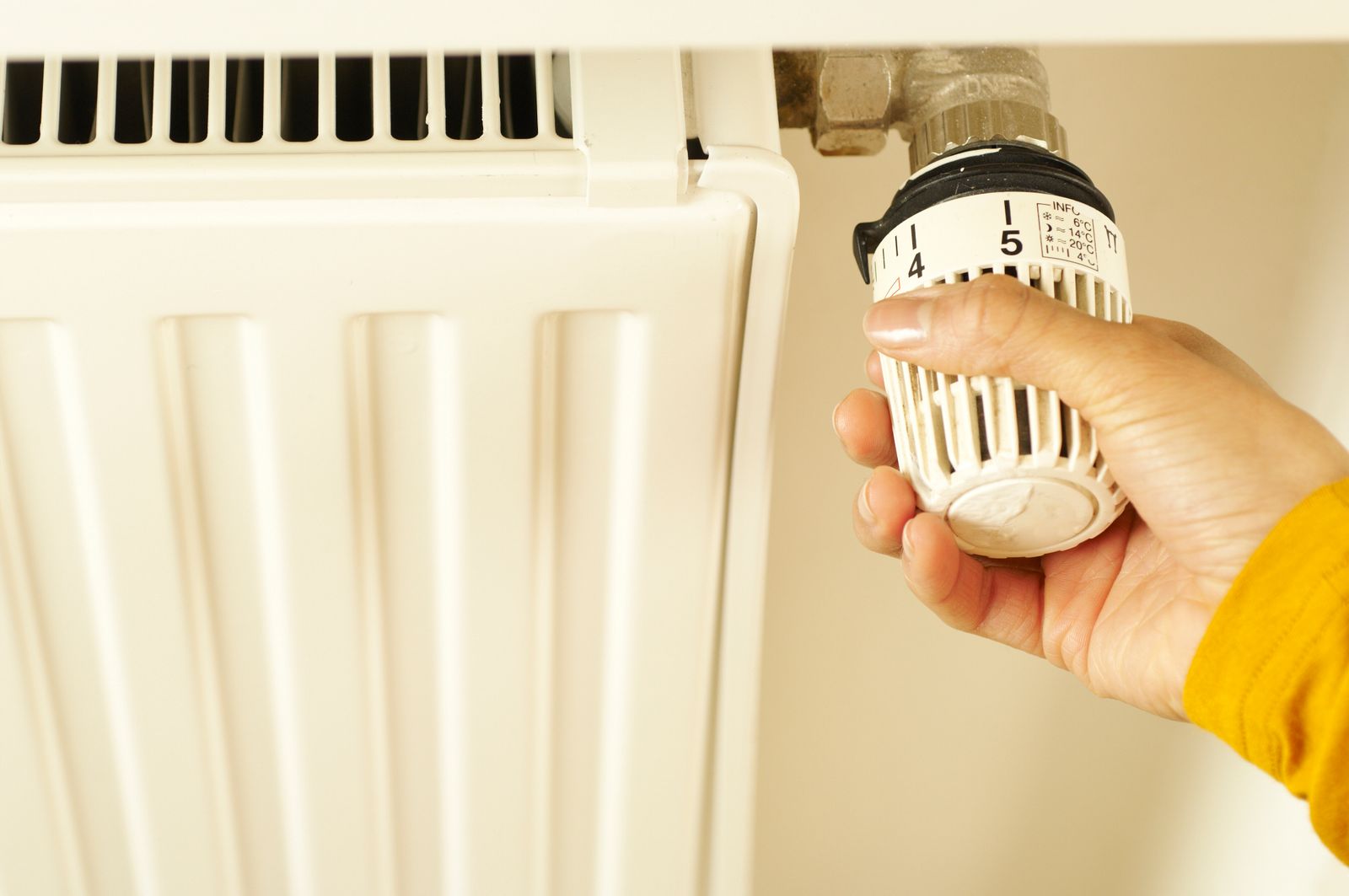 A hand in a yellow sleeve turning the thermostatic valve on a white panel radiator