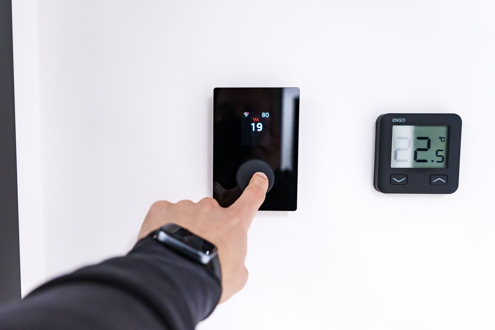 A finger pressing a black wall thermostat set to 19 degrees, with a second small thermostat beside it
